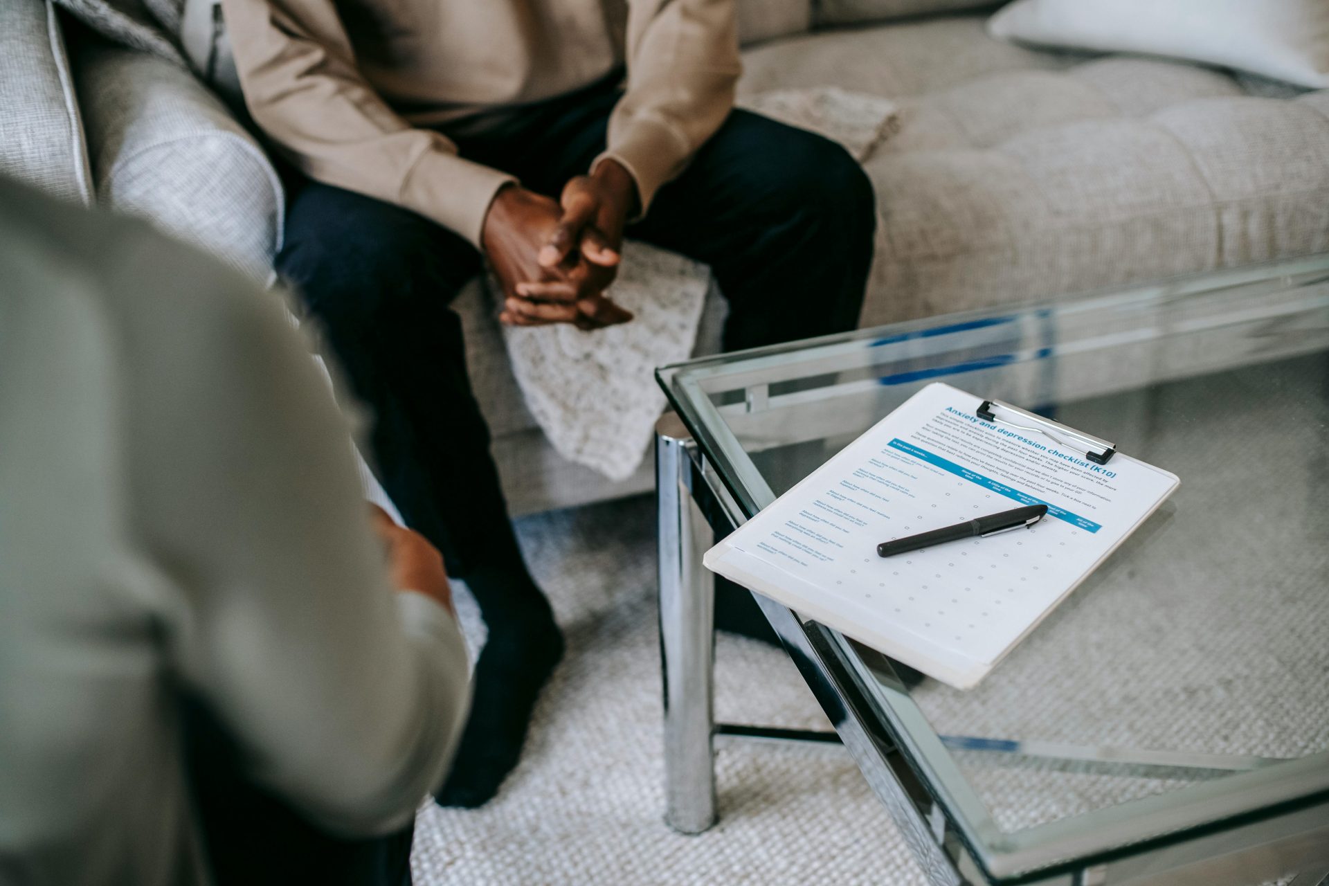 From above of unrecognizable ethnic male patient with clasped hands sitting near anonymous psychologist in office with glass table and clipboard with notes